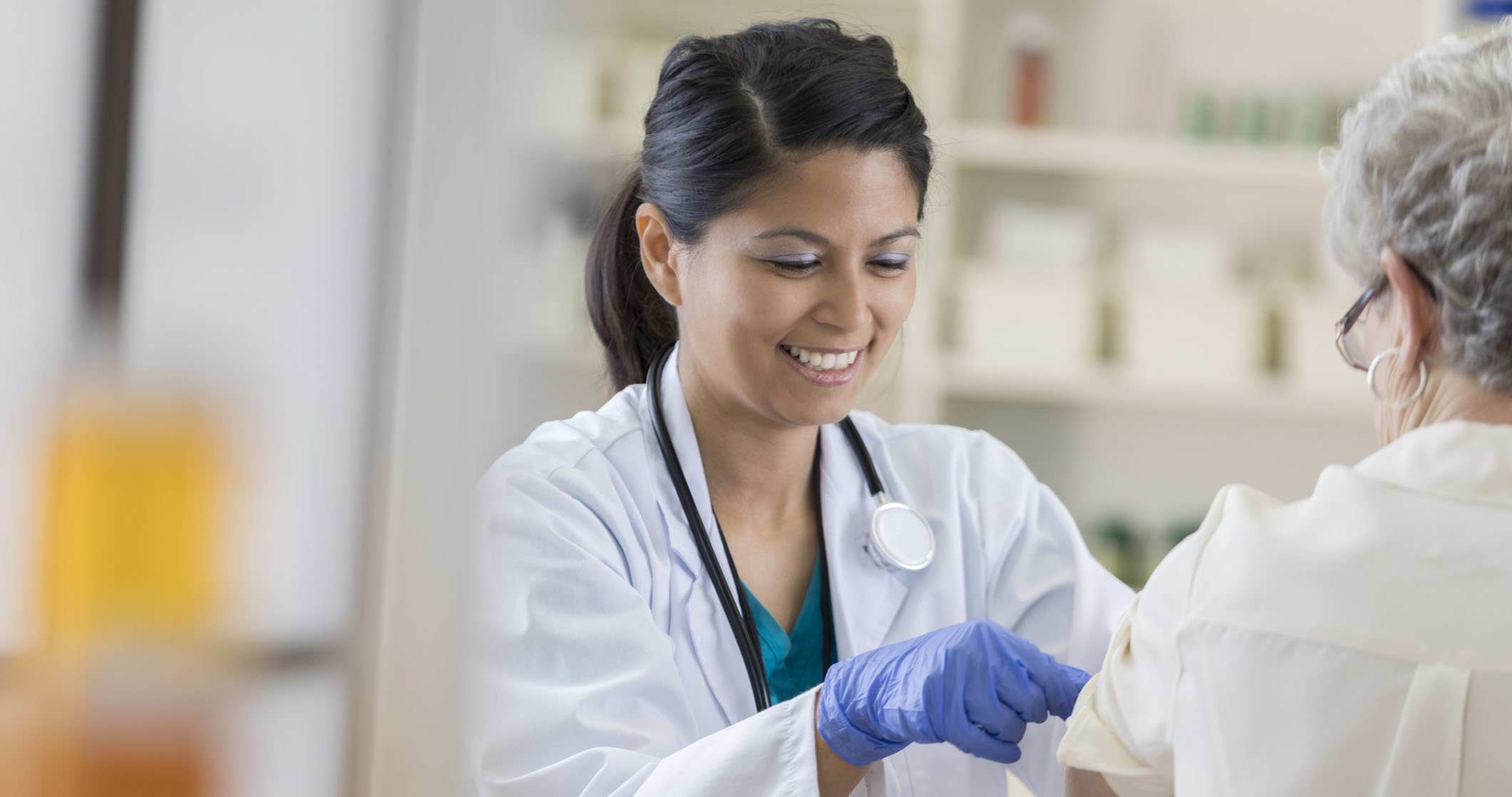 A smiling young pharmacist dons a stethoscope and completes her senior customer's flu shot by placing a band-aid on her upper arm.  She is wearing medical gloves and a lab coat.  There are shelves in the background.