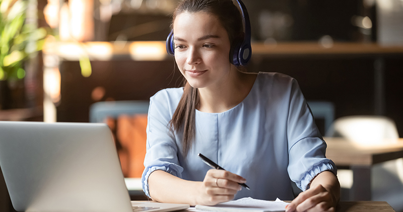 Focused woman wearing headphones using laptop, writing notes